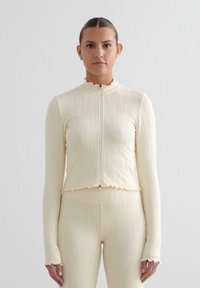 Top lungo a maniche ribbed color crema con zip frontale, colletto ondulato e polsini. Il tessuto è testurizzato, il design aderente mette in risalto la silhouette.