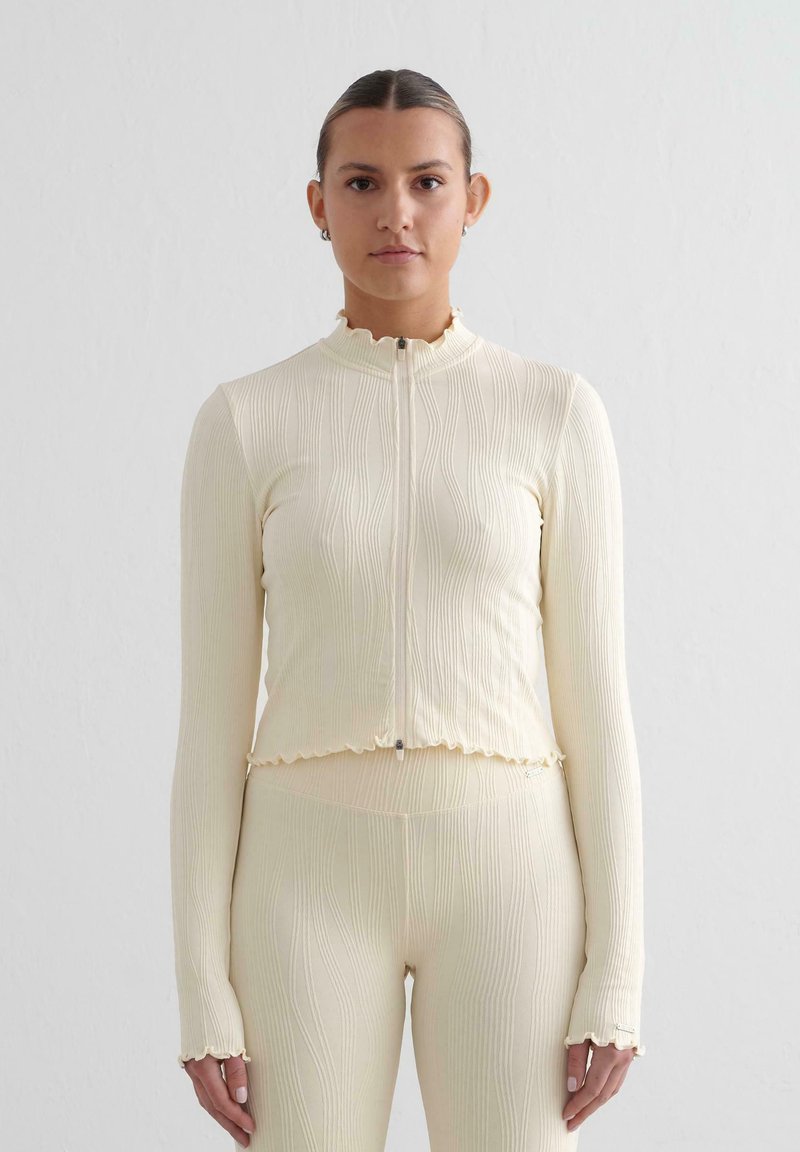 Top lungo a maniche ribbed color crema con zip frontale, colletto ondulato e polsini. Il tessuto è testurizzato, il design aderente mette in risalto la silhouette.