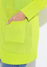 Maglione verde lime con un design a rete testurizzata, con tasca frontale e polsini a costine. Il tessuto testurizzato migliora la traspirabilità.