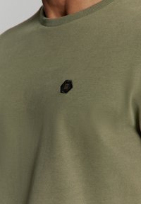 Olijfgroene sweatshirt gemaakt van zachte stof, met een geribbelde ronde hals en een klein zwart hexagonaal logo op de borst.