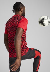 Rdeča športna majica z črnim geometrijskim vzorcem, v kombinaciji s črnimi športnimi hlačami, ki imajo rdeče bočne akcentne. Nogometna žoga v gibanju.