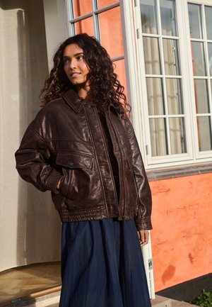 Femme aux cheveux bouclés portant une veste en cuir marron foncé et une longue jupe bleu marine, debout à l'extérieur près d'un mur couleur pêche avec des fenêtres aux cadres blancs.