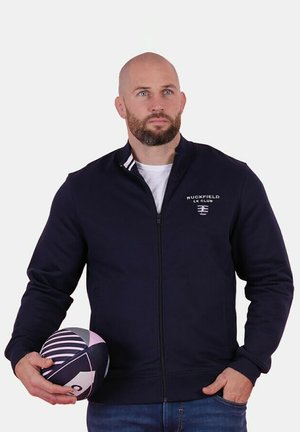 Homme portant une veste zippée bleue marine, tenant un ballon de rugby rayé d'une main, regardant légèrement sur le côté contre un fond uni.