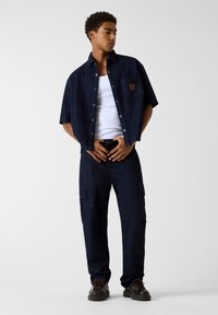 Ciemnoniebieska koszula z krótkim rękawem i kieszenią na piersi, w połączeniu z ciemnymi spodniami typu cargo z bocznymi kieszeniami, noszona z brązowymi sznurowanymi butami.