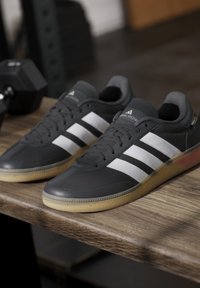 Black athletic shoes with three white stripes, synthetic material, flat rubber sole with light brown accents, and gray padded collar.