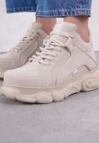 Beige sneakers met een bovenwerk van soepel leer, een dikke gestructureerde zool en een ronde neus. Voorzien van een vetersysteem en branding op de zijkant.