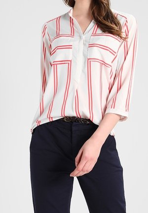 Frau trägt eine weiße Bluse mit roten vertikalen und horizontalen Streifen sowie eine dunkelblaue Hose mit einem schwarzen geflochtenen Gürtel.