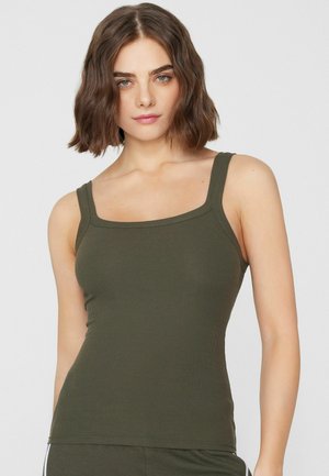 Jeune femme aux cheveux bruns courts et ondulés portant un débardeur côtelé vert olive sur un fond clair uni.