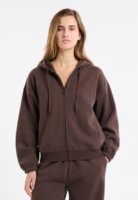 Brązowa bluza z kapturem na zamek, o luźnym kroju, z elastycznymi mankietami i gładką fakturą. W komplecie z dopasowanymi spodniami w podobnym kolorze.
