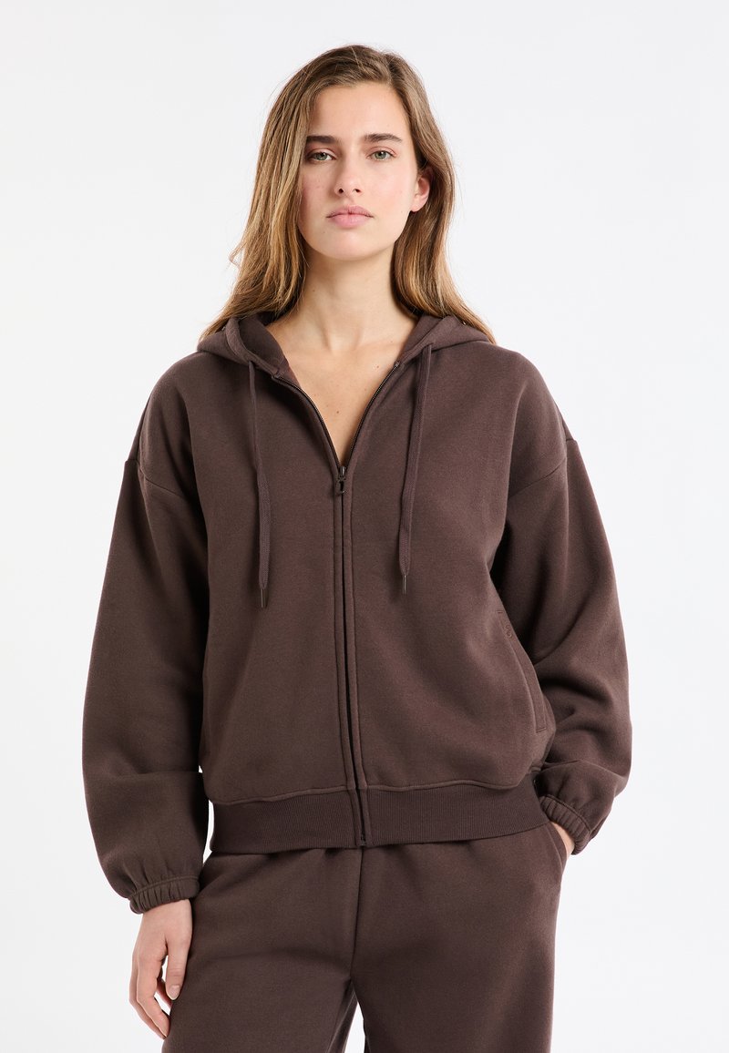 Brązowa bluza z kapturem na zamek, o luźnym kroju, z elastycznymi mankietami i gładką fakturą. W komplecie z dopasowanymi spodniami w podobnym kolorze.