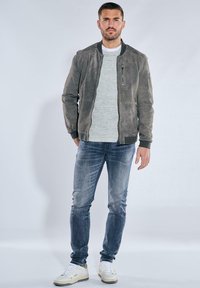 Graue Wildleder-Bomberjacke mit gerippten Bündchen, getragen über einem hellgrauen Pullover, kombiniert mit blauen Slim-Fit-Jeans und weißen Sneakers.