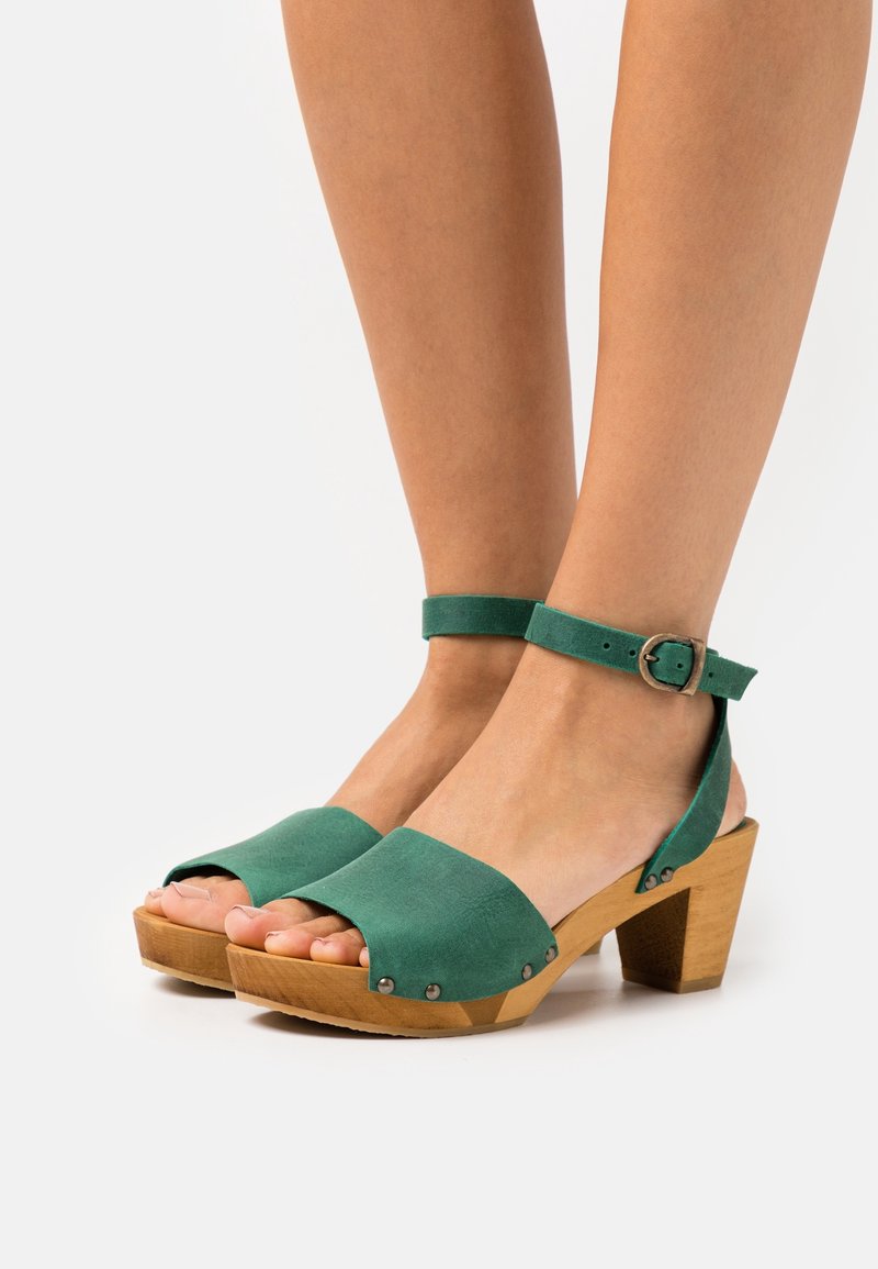 Jambes portant des sandales ouvertes en daim vert avec talons blocs en bois et brides de cheville avec boucles métalliques.