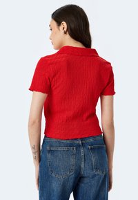Top corto a maniche corte rosso con texture e colletto, abbinato a jeans di denim blu con tasche posteriori; il modello è mostrato di spalle.