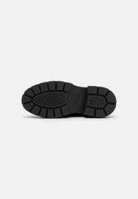 Suela de zapato de goma negra con un patrón de textura, que presenta surcos y crestas profundas para mayor tracción. Marcada "GUESS" en el costado.