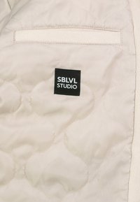 Beige gesteppte Jacke mit kreisförmigen Stickmustern, einem genähten schwarzen Etikett mit der Aufschrift "SBLVL STUDIO" und einer versteckten Tasche mit Klettverschluss.