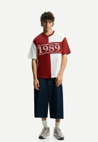 T-shirt bloccato in rosso e bianco con "THE VICTORIOUS ARENA 1989" stampato sul davanti; abbinato a pantaloni shorts larghi in denim scuro e sneakers.