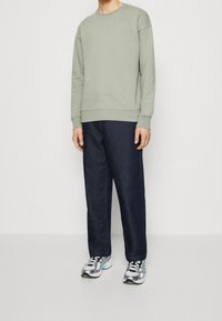 Lichtgroene trui met ronde hals en geribbelde boorden, gecombineerd met donkerblauwe wijde jeans. Schoeisel bestaat uit grijze en zwarte sportschoenen.