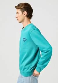Sweatshirt azzurro chiaro a maniche lunghe con il logo ricamato "Wrangler Jeans". Presenta polsini e orlo a coste. Materiale morbido, vestibilità comoda.