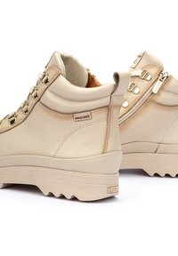 Beige leather ankle boots with metal eyelets and zipper, textured upper, chunky rubber sole, logo detail on the side.
