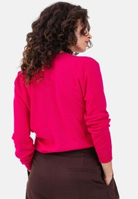 Fuchsiafarbener Strickpullover mit langen Ärmeln, gerippten Bündchen und rundem Ausschnitt, kombiniert mit dunkelbraunen Hosen mit Gesäßtaschen.