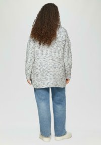Lange mouwen, oversized trui in een grijs en off-white gespikkeld patroon, gecombineerd met lichtblauwe jeans en witte platform sneakers.
