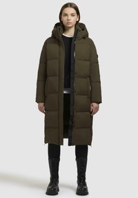 Olive green padded coat with a hood, featuring a zip closure, side pockets, and a long design, paired with black leggings and combat boots.