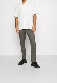 Biała koszula z krótkim rękawem i kołnierzykiem, z kieszenią na piersi, zestawiona z brązowymi spodniami w kratę i czarnymi butami na sznurówki z grubą podeszwą.