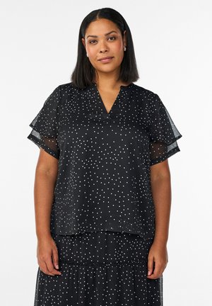 Ženska s črnimi srednje dolgimi lasmi, oblečena v črno obleko s kratkimi rokavi in belimi pikami na belem ozadju.