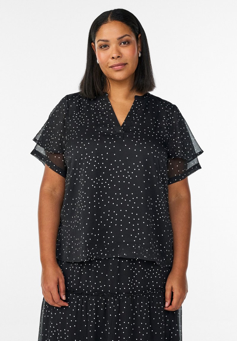 Kobieta o średniej długości czarnych włosach, ubrana w czarną sukienkę z krótkim rękawem w białe kropki na białym tle.