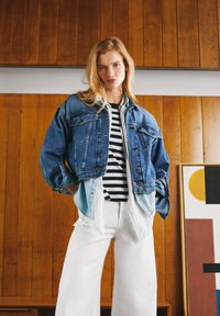 Veste en denim bleu raccourcie avec des poches poitrine, superposée sur une chemise rayée noire et blanche, assortie avec des pantalons larges blancs.