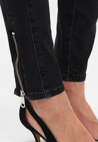 Jeans de mezclilla negra con un detalle de cremallera en el tobillo. Tienen un corte ajustado. Llevados con zapatos de tacón negros.