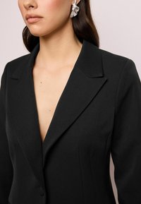 Schwarzer Blazer mit taillierter Passform, Reverskragen und tiefem V-Ausschnitt. Der Stoff wirkt glatt. Ohrringe in Blattform aus Silber runden den Look ab.
