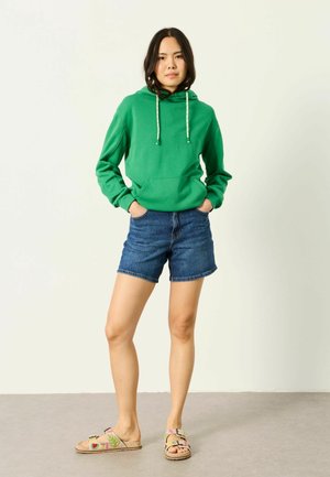 Junge Frau steht mit den Händen in den Taschen, trägt einen grünen Hoodie, Jeansshorts und Sandalen mit Blumenmuster vor einem einfarbigen Hintergrund.