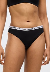 Čierne bavlnené tangá s bielym elastickým pásom, na ktorom je čiernym tlačeným nápis "KARL LAGERFELD". Hladká textúra a minimalistický dizajn.