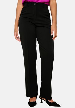 Kobieta ubrana w czarne spodnie o prostych nogawkach i wysokim stanie, połączone z czarnymi sandałkami na obcasie z odkrytą piętą i wsuniętą do środka różową bluzką.