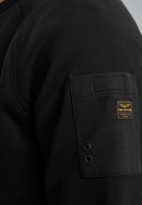 Sudadera negra con tejido texturizado, un bolsillo en la manga con una etiqueta amarilla y dos ojales reforzados.