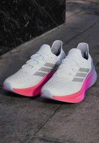 Scarpe da corsa bianche con una tomaia testurizzata, dotate di righe grigie e una suola con gradiente rosa brillante. Design leggero con colletto imbottito.