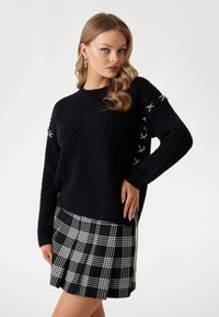 Zwarte gebreide trui met een gestructureerd patroon en geborduurde accenten op de mouwen, gecombineerd met een geplooide zwart-witte geruite rok.