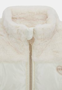 Giacca imbottita di colore chiaro con un colletto morbido, zip dorata e una patch a forma di cuore. Texture morbida con materiali a contrasto.