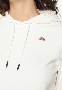 Sudadera blanca hecha de tejido suave, con un bolsillo tipo canguro en la parte delantera, cordón ajustable y un pequeño logo marrón en el pecho.