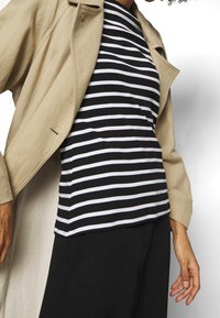 Persona che indossa un cappotto beige sopra una camicia a righe bianche e nere con pantaloni neri, in piedi su uno sfondo bianco.