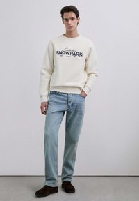 Sudadera crema con grafismo "SNOWPARK", combinada con vaqueros azul claro. Corte ajustado, textura suave. Botas marrones resistentes completan el conjunto.