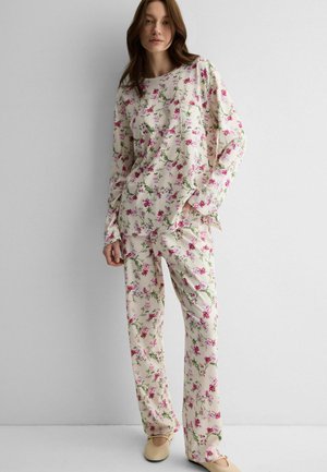 Donna che indossa una maglia a maniche lunghe color crema e pantaloni con stampa floreale rosa e verde, in piedi contro una parete bianca, scarpe beige.