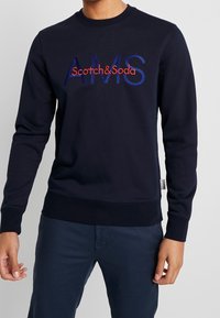 Sudadera navy con cuello redondo, puños y dobladillo acanalados. Presenta el logo de "Scotch&Soda" y "AMS" bordados en rojo y azul. Textura de tela suave.