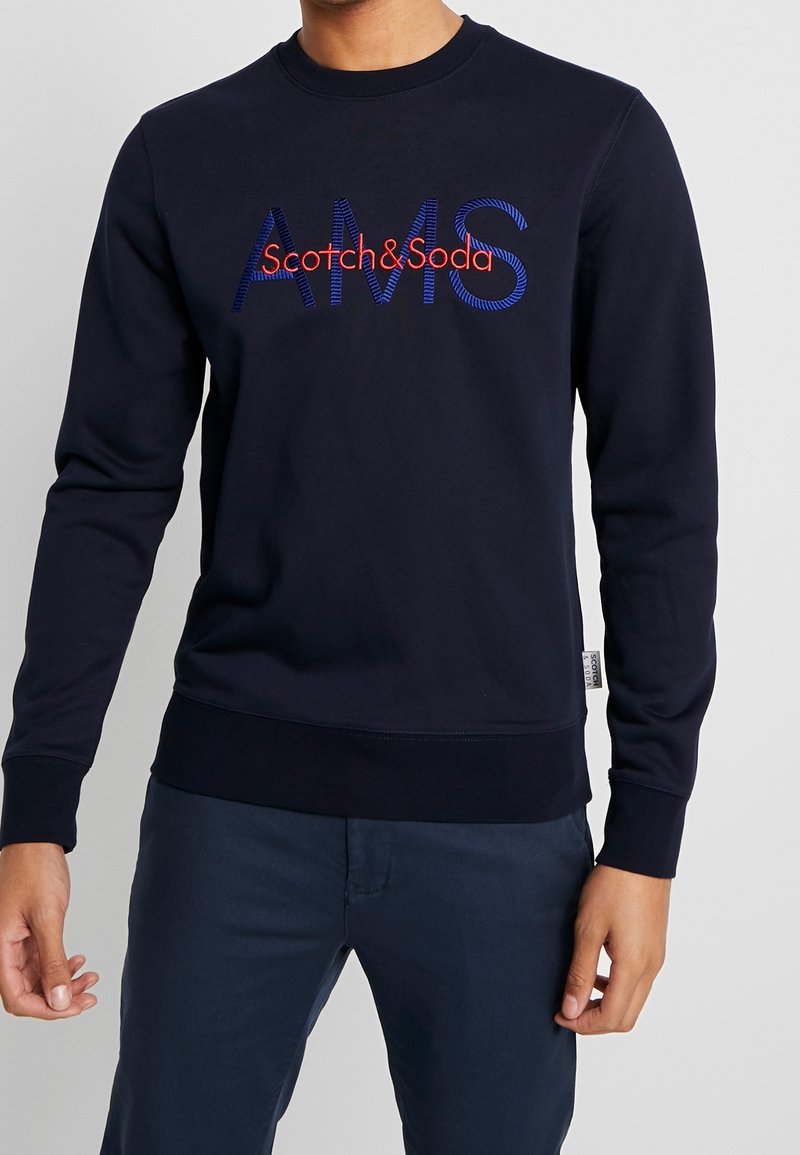 Sudadera navy con cuello redondo, puños y dobladillo acanalados. Presenta el logo de "Scotch&Soda" y "AMS" bordados en rojo y azul. Textura de tela suave.