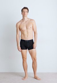 Črne bokserice z barvitim pikčastim vzorcem, prilagojene zasnove in mehak elastičen pas. Model stoji bos na svetli podlagi.