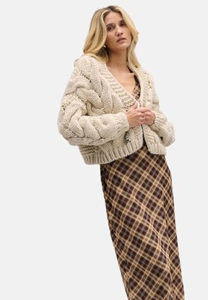 Gruba kremowa rozpinana kardigan z dzianiny warkoczowej o skróconym kroju; zestawiona z długą brązową sukienką w kratę z dekoltem w serek i subtelną fakturą.