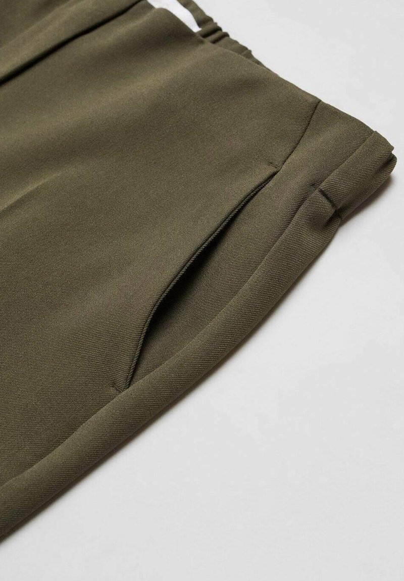 Primo piano di pantaloni verde oliva con tasca laterale e cintura elastica su uno sfondo chiaro.