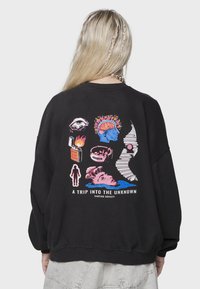 Sudadera negra con gráficos coloridos en la parte posterior, que incluyen ojos, un cerebro y diseños abstractos, con el texto "UN VIAJE AL DESCONOCIDO."