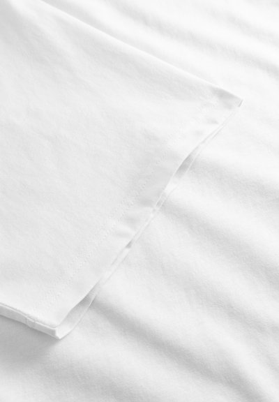 Κοντινό πλάνο μανικιού λευκού T-shirt με εμφανείς ραφές σε λείο φόντο από ύφασμα.